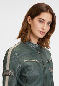 Giacca da moto in pelle verde con colletto alto, due tasche frontali con zip, strisce beige sulle spalle e toppe testurizzate.