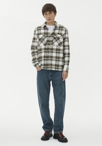 Młody mężczyzna stoi, ubrany w biało-zieloną koszulę w kratę z guzikami, niebieskie dżinsy i czarne buty z czerwonymi sznurówkami, częściowo trzymając ręce w kieszeniach.