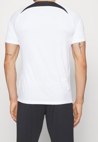 Biała sportowa koszulka z krótkim rękawem, z czarnym akcentem na plecach oraz gładką, syntetyczną fakturą. Dopasowany krój z bocznymi szwami.