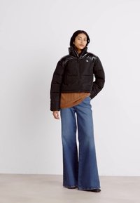 Zwarte cropped pufferjack met glanzende schouders, rits aan de voorkant en logo. Draag het over een bruine geribbelde trui, gecombineerd met flared blauwe jeans.