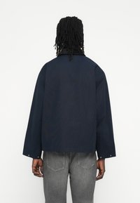 Veste bleu marine à coupe décontractée, épaules tombantes et col côtelé. Dispose de pressions aux poignets et d'un tissu lisse et texturé.