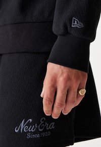 Schwarzer ribbeliger Pullover mit umgeschlagenen Ärmeln, mit einem gestickten "New Era Since 1920"-Logo und einem goldenen Ring an einer Hand.