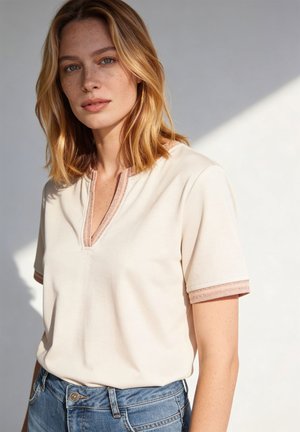 Blondynka z piegami nosi beżowy top z krótkim rękawem i koronkowym obszyciem oraz niebieskie dżinsy, stoi na tle neutralnego, jasnego tła.