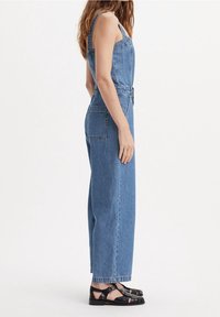 Ljusblå denimoverall med vida ben, justerbara axelremmar och sidfickor, i kombination med svarta sandaler med remmar. Slät yta.