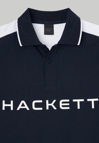 Námornícky modrá polo tričko vyrobené z textúrovanej tkaniny, s bielym golierom a výrazným bielym logom "HACKETT" na prednej strane.