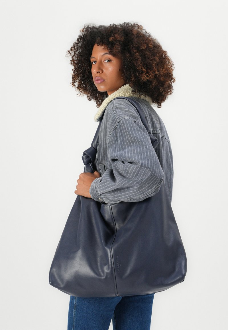 Navyblaue Leder-Totetasche mit einem lässigen Design, ausgestattet mit einer glatten Textur und einem Knotenakzent am Trageriemen, gehalten von einer Person.