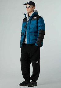 Giacca a vento blu e nera con cuciture orizzontali, felpa grigia, pantaloni in fleece neri e calzature nere. Include occhiali da sole e cappellino.