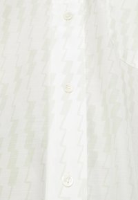 Chemise blanche avec un motif en zigzag subtil de couleur vert clair, dotée d'une patte de boutonnage et de trois boutons à l'avant. Texture de tissu douce.