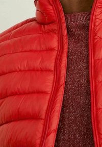 Veste matelassée rouge avec un col montant, dotée de surpiqûres matelassées et d'une fermeture éclair frontale. Portée par-dessus un pull texturé rouge foncé.