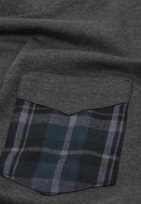 Jersey gris con un bolsillo con parche que presenta un estampado a cuadros en azul y negro. Tejido suave y texturizado con un detalle en el borde del bolsillo en corte de espiga.