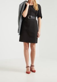 Schwarzes Kurzarmkleid mit taillierter Silhouette, betont an der Taille mit einem strukturierten Gürtel, kombiniert mit roten Wildleder-Ankelriemchen-Absätzen und einem grauen karoartigen Blazer.