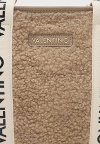 Bež faux karusnahk kott tekstuuriga pinnaga. Omab logo plaastrit ja kanvasrihme, millel on musta värviga trükitud "VALENTINO". Puhtad jooned ja minimaalne disain.