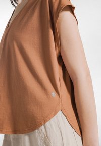 Brun bomulds V-hals top med korte ærmer, der har en rundet kant og et lille logo på siden; båret over et lyst stof.