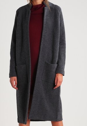 Femme portant un cardigan gris foncé ouvert, à manches longues et longueur genou, par-dessus une robe bordeaux, sur un fond clair uni.