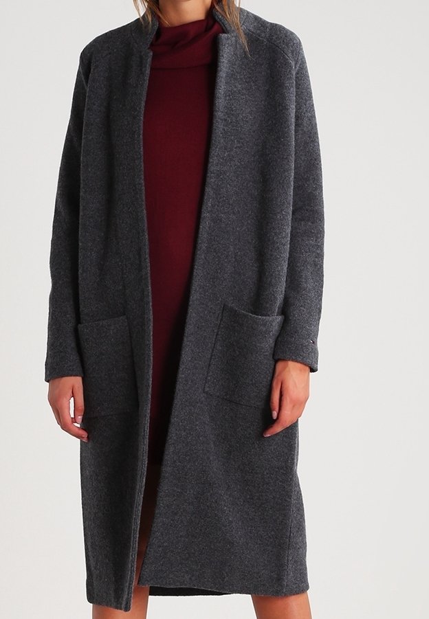 Femme portant un cardigan gris foncé ouvert, à manches longues et longueur genou, par-dessus une robe bordeaux, sur un fond clair uni.
