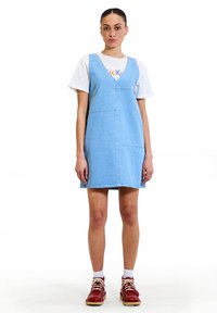 Abito in denim blu chiaro con scollo a V, tasche frontali e dettagli di cucitura, abbinato a una t-shirt bianca e scarpe rosse con lacci.