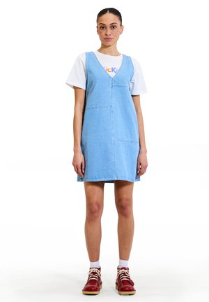Vestido de mezclilla azul claro con escote en V, bolsillos frontales y detalles de costura, combinado con una camiseta blanca y zapatos rojos con cordones.