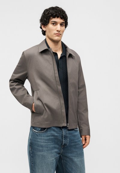 Νέος άνδρας με σγουρά σκούρα μαλλιά, φοράει γκρι ζακέτα με φερμουάρ πάνω από μαύρο μπλουζάκι και μπλε τζιν, στέκεται με το ένα χέρι στην τσέπη.