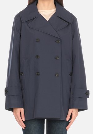 Kvinde iført navyblå dobbeltradet frakke med brede slag og knapmanchetter, kombineret med blå jeans og beige top under.