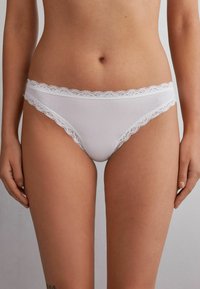 Culotte en coton blanc avec bordure en dentelle à la taille et aux ouvertures des jambes. Présente une texture lisse et une coupe taille basse pour un confort optimal.