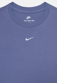 Nike majica v modrem bombažu, ki ima rebrast krojeni izrez in bel Nike swoosh logotip na prsih. Gladka tekstura z minimalnimi podrobnostmi.