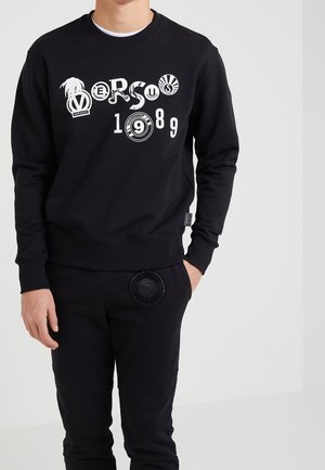Ein Mann trägt einen schwarzen Pullover mit weißem "VERSUS 1989" Schriftzug und schwarze Hosen mit rundem Emblem auf der Tasche, steht vor einer weißen Wand.