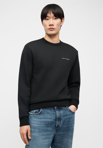 Νεαρός άνδρας που φοράει μαύρο φούτερ με στρογγυλή λαιμόκοψη και το κείμενο "Tiger of Sweden" και μπλε τζιν, στέκεται με το ένα χέρι στην τσέπη.