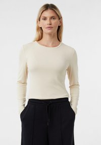 Jasnobeżowa bluzka z długim rękawem o okrągłym dekolcie i dopasowanym kroju. Miękka, teksturowana tkanina zestawiona z czarnymi spodniami ze ściągaczem.