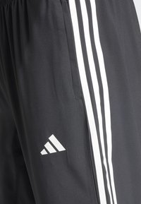 Schwarze Sporthose aus glattem, leichtem Stoff. Mit drei weißen vertikalen Streifen an einer Seite und einem weißen Logo auf dem Oberschenkel.