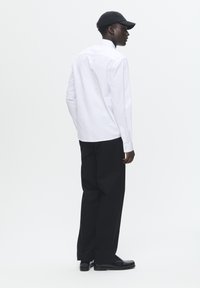 Camisa blanca de manga larga con puños abotonados, combinada con pantalones negros y zapatos negros. Fondo blanco liso. La gorra añade un detalle elegante.