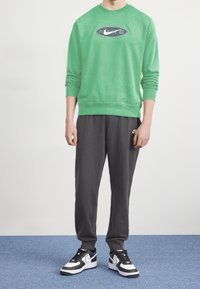 Grön sweatshirt med en logotyp, parat med mörkgrå sweatpants. Svart och vita sneakers fulländar outfiten. Avslappnad atletisk stil.