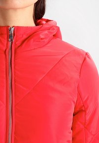 Rode gewatteerde jas met capuchon, voorzien van een ritssluiting aan de voorkant, een doorgestikte textuur, lichtgewicht materiaal, zichtbare stiknaden en een gladde afwerking.