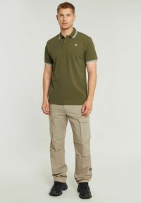 Polo verde oliva con accenti a righe bianche, abbinato a pantaloni cargo beige con tasche e calzature nere.
