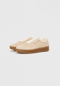 Beżowe zamszowe sneakersy z teksturowaną gumową podeszwą, wyposażone w kremowe sznurówki i miękką wyściółkę wewnętrzną. Prosty design z minimalnymi akcentami.