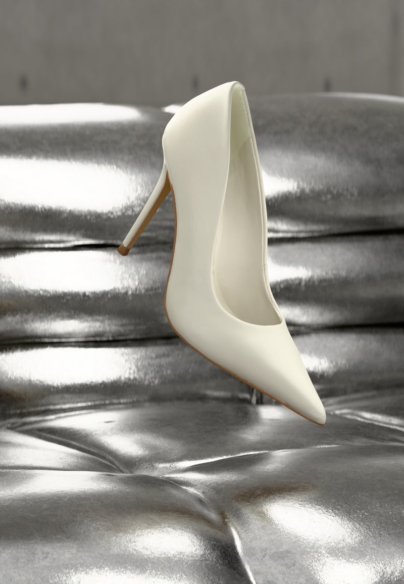 Escarpin blanc à talon haut avec un bout pointu, en cuir lisse, et une semelle beige, posé sur une surface métallique texturée.