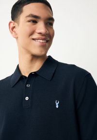 Marineblå polo-shirt med klassisk krave, to knapper og en lyseblå broderet logo på brystet. Standard pasform og tekstureret stof.