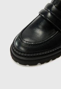 Black leather loafer featuring a rounded toe, decorative stitching, two upper straps, and a thick rubber sole with a textured tread pattern.