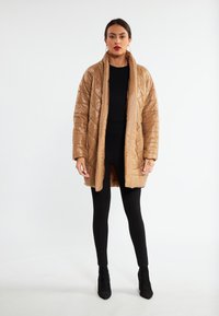 Tan prošívaný puffer kabát s jemným leskem, oversized designem, širokým límcem a bočními kapsami, nošený přes černý top a legíny.
