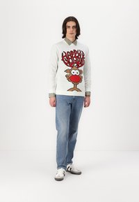 Suéter blanco con un gráfico festivo de reno, detalles en rojo y el texto "Felices Fiestas". Combinado con jeans azules y zapatillas blancas.