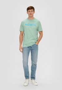 Mintgrünes Baumwoll-T-Shirt mit einem blauen Grafikdruck, kombiniert mit hellblauen Jeans und weißen Turnschuhen, vor einem neutralen Hintergrund stehend.