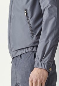 Szara, lekka kurtka z zamkiem błyskawicznym na piersi, elastycznym paskiem w talii i ściągaczami na mankietach; gładka tekstura i elegancki, nowoczesny design.