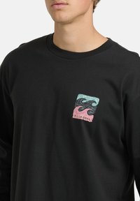 Junger Mann trägt ein schwarzes Langarmshirt mit einem kleinen, bunten Billabong-Logo, das Wellenmuster auf der Brust zeigt.