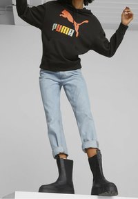 Musta huppari, jossa on suuri oranssi PUMA-logo, vaaleansiniset käärityt farkut ja mustat paksupohjaiset nilkkurit.