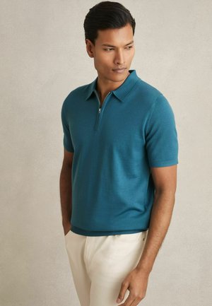 Teal kortærmet polo shirt med zipkrave, lavet af blødt stof, parret med lysfarvede bukser. Glat tekstur og rene linjer.