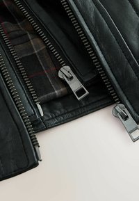 Zwarte leren jas met meerdere metalen ritsen. Interieur heeft een ruitpatroon in donkere tinten met rode accenten. Getextureerd lederen oppervlak.