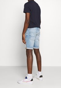 Hombre con pantalones cortos de mezclilla azul claro, camiseta navy, calcetines blancos y zapatillas Nike blancas de pie de espaldas sobre un fondo liso.