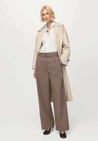 Béžový trenčkot cez bielu košeľu, doplnený hnedými širokými nohavicami s vysokým pásom. Čierne lodičky dokončujú vzhľad.