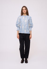 Blusa azzurro chiaro con motivo a pallini e maniche a sbuffo, caratterizzata da design floreali e geometrici; abbinata a pantaloni scuri e larghi e scarpe nere.