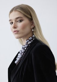 Lyshåret kvinde iført sort fløjlsjakke, stor metallisk sølvkugle-halskæde, matchende hængende øreringe og neutral makeup mod hvid baggrund.