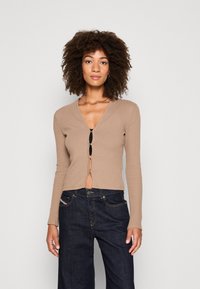 Beige ribbed cardigan with a V-neck and button closure, featuring long sleeves and a cropped design, paired with dark blue jeans.